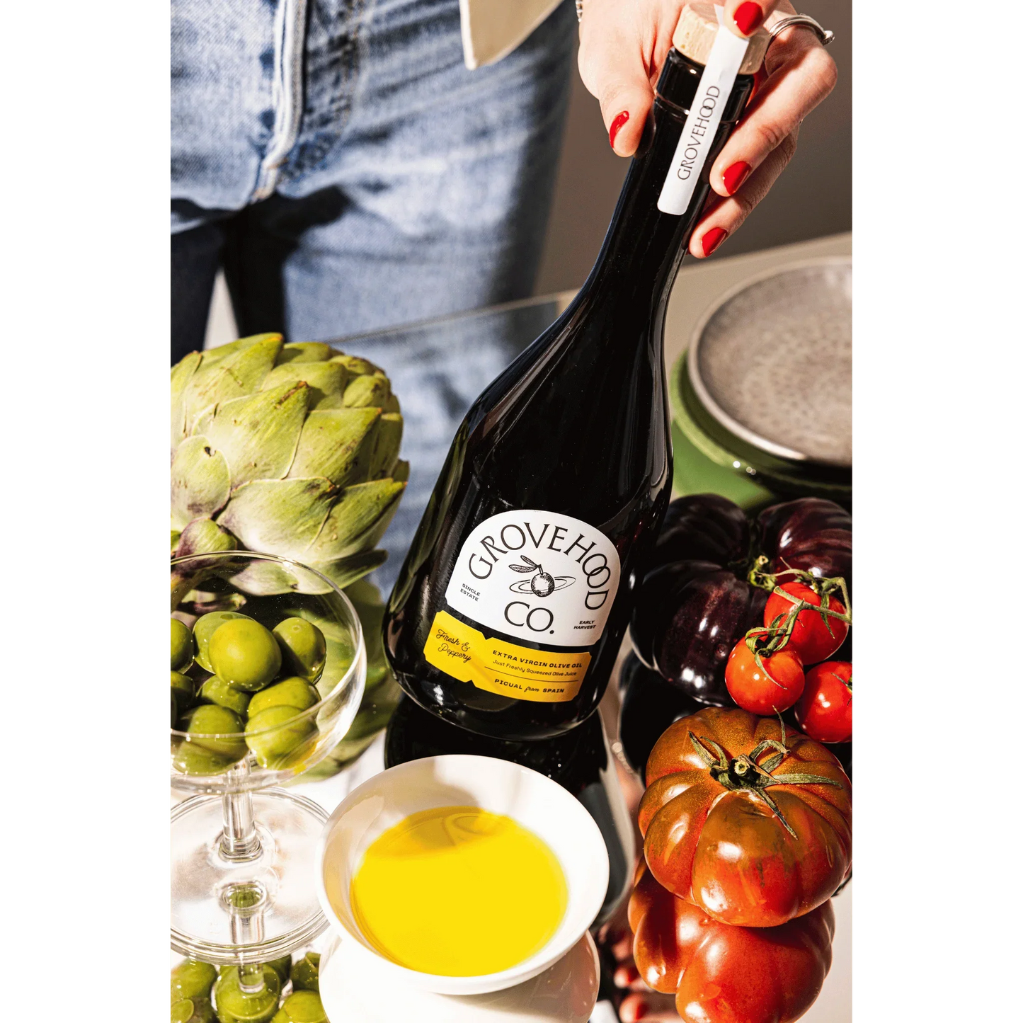 Grovehood Collective Spanish extra virgin olive oil bottle at a table with tomatoes, olives and artichoke