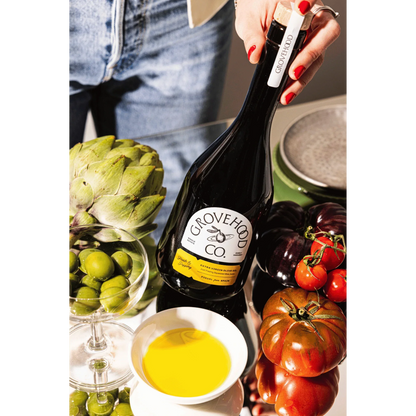 Grovehood Collective Spanish extra virgin olive oil bottle at a table with tomatoes, olives and artichoke