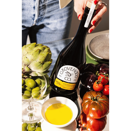 Grovehood Collective Spanish extra virgin olive oil bottle at a table with tomatoes, olives and artichoke